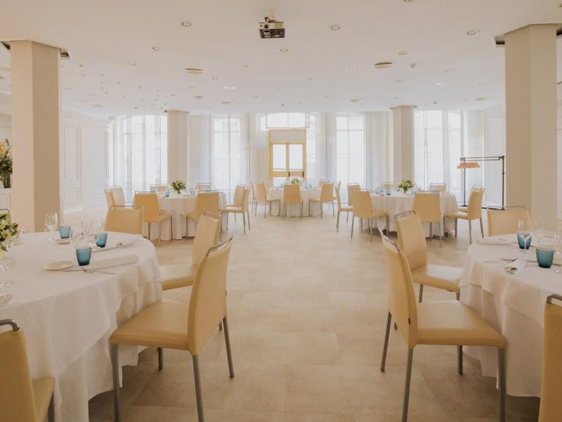 Marina Room bright event space at Lamaro Hotel with round tables dressed in crisp white linens and beige chairs, accented by fresh floral centerpieces and vibrant turquoise glassware.