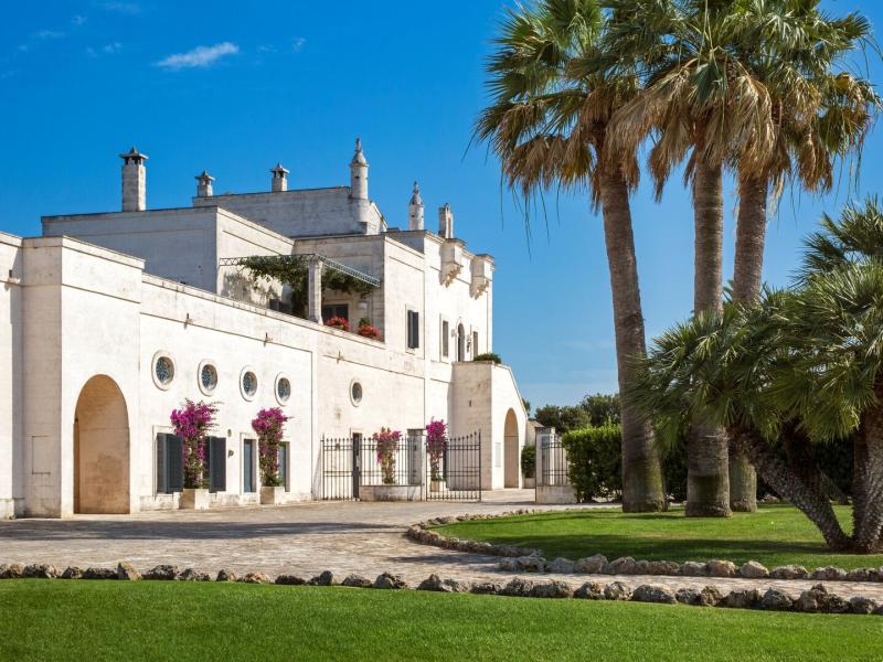 Masseria San Domenico stunning whitewashed exterior framed by towering palm trees and vibrant magenta bougainvillea, set against a bright blue sky and lush green lawn.