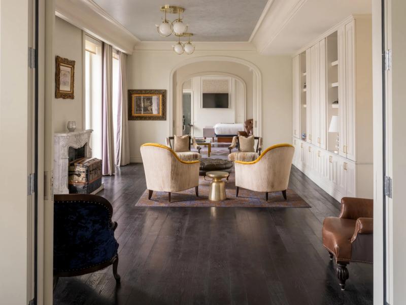 The grand living space of the The Owners Box Suite features dark hardwood floors and cream-paneled walls, complemented by golden armchairs and deep blue accents for a refined, inviting atmosphere.