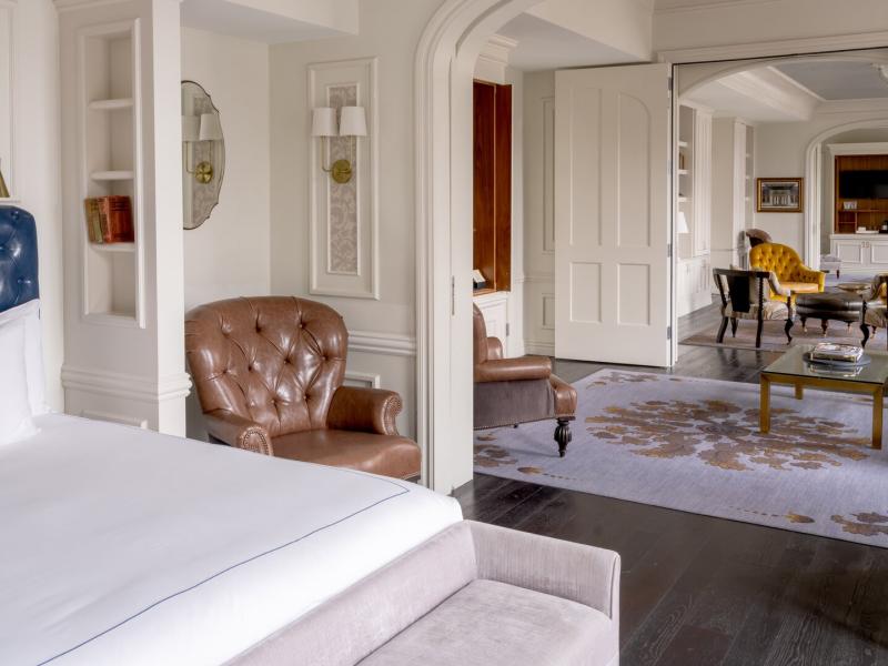 The Owners Box Suite showcasing a luxurious suite with a tufted navy headboard and plush brown leather chair, accented by soft ivory walls and a patterned gray rug for an elegant retreat.