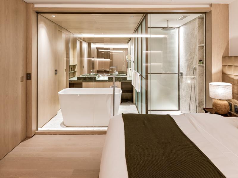 A serene suite bedroom with light oak walls and an olive-green bed throw, opening to a spa-like bathroom with marble accents and a freestanding tub.