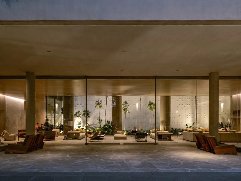 Pulso Hotel grand entrance floor-to-ceiling glass panels paired with warm taupe columns create a serene, light-filled entrance accented by verdant foliage and ambient golden lighting.