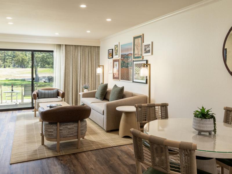 A spacious living space in a Premium Golf View 1 Bedroom King Suite with cozy seating and dining areas, accented by earthy brown chairs, soft green pillows, and warm wood flooring.