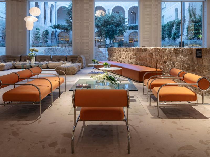 The Jaffa Hotel's modern lobby lounge area features sleek orange seating and glass tables, complemented by natural stone textures and soft beige tones.