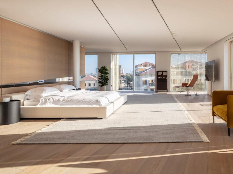 Spacious Penthouse bedroom with floor-to-ceiling windows and sleek wood paneling, accented by mustard armchairs and soft beige tones.