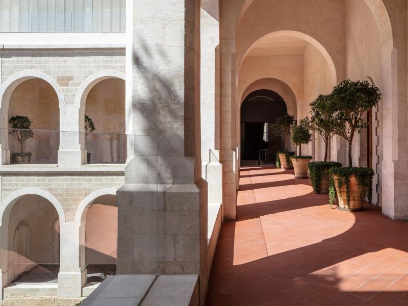 A sunlit corridor leading to guest accommodations is lined with terracotta flooring and potted greenery, contrasted against crisp white arches and soft shadow play.