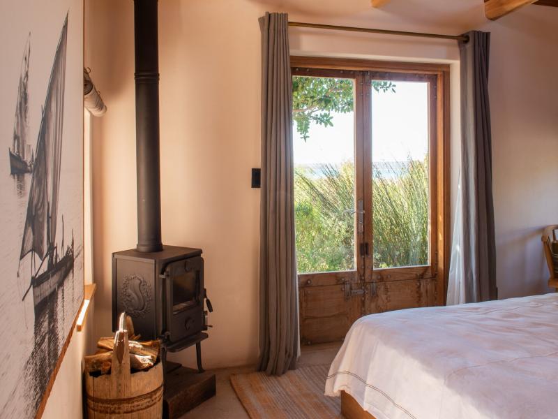 Yellowwood Cottage bedroom features warm and inviting atmosphere with soft beige walls accented by natural wood tones, featuring a cozy wood-burning stove and large glass doors framing lush outdoor greenery.