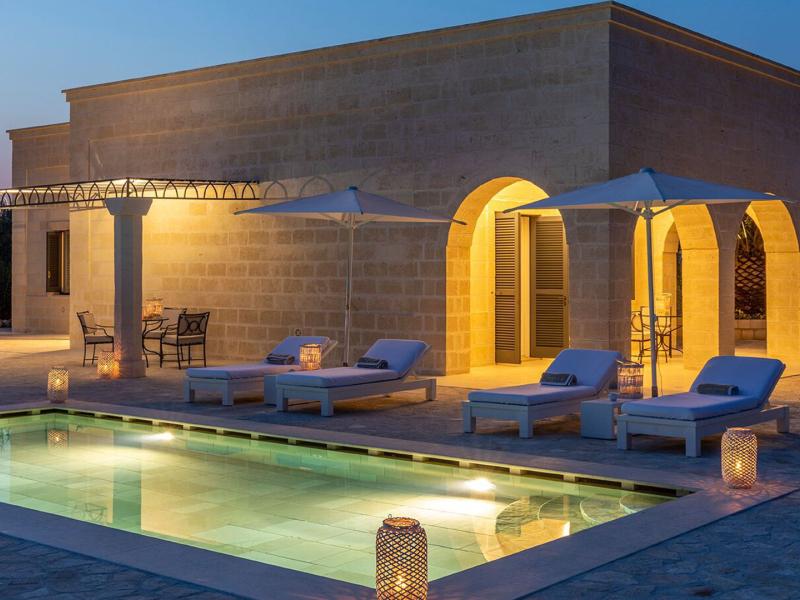 A serene stone villa, the Villino (Villa) di San Domenico illuminated by warm golden lights, featuring a sparkling turquoise pool and soft beige accents under a twilight sky.