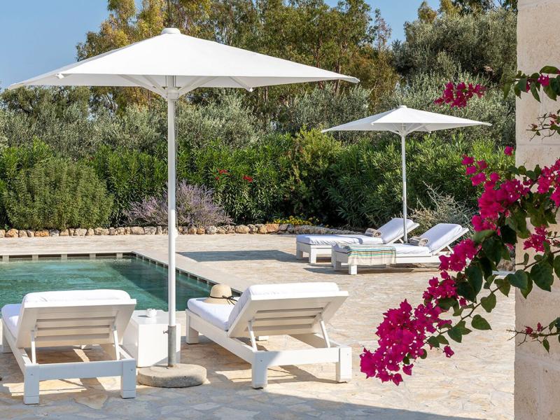 The Villino (Villa) di San Domenico sunlit terrace with crisp white loungers and umbrellas beside a deep green pool, framed by lush greenery and vibrant pink bougainvillea.