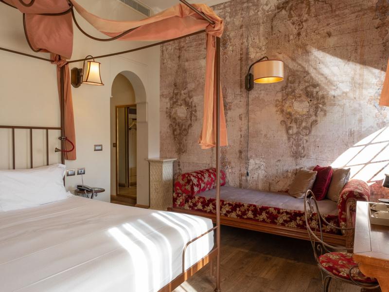 Hotel Ville sull'Arno Executive Triple guestroom with stylish canopy bed with warm terracotta drapes and rustic wood flooring pairs beautifully with a textured accent wall and deep red details.