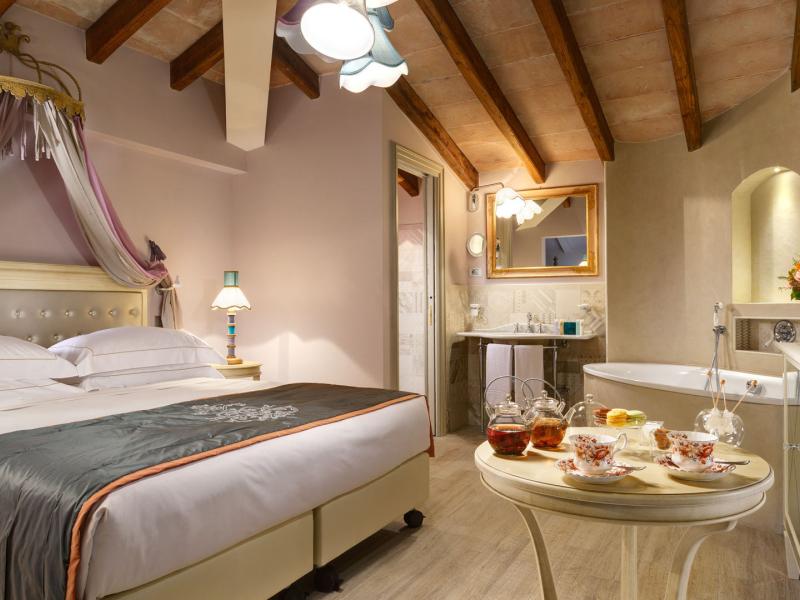 Hotel Ville sull'Arno Junior Suite with a charming canopy, flowing lavender and cream fabrics complements exposed wooden beams and a chic sitting area, accented by teal and blush tones.