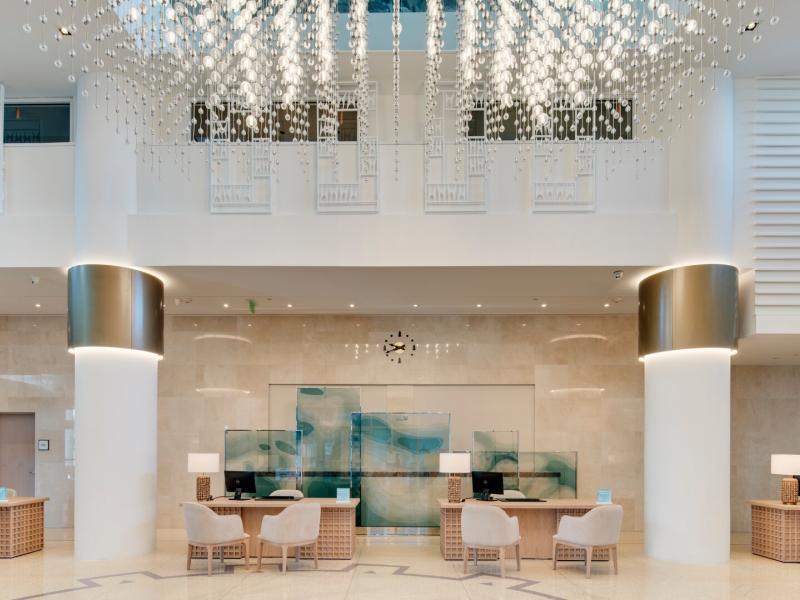 Carillon Miami Wellness Resort bright, airy lobby with chic beige and white tones, accented by a stunning crystal chandelier and calming teal artwork.