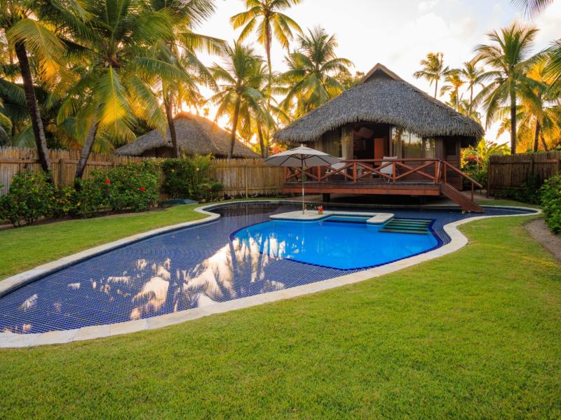 A serene Garden Bungalow exterior with a sleek blue pool framed by vivid green grass and tropical palms, complemented by warm wooden accents and soft beige tones.