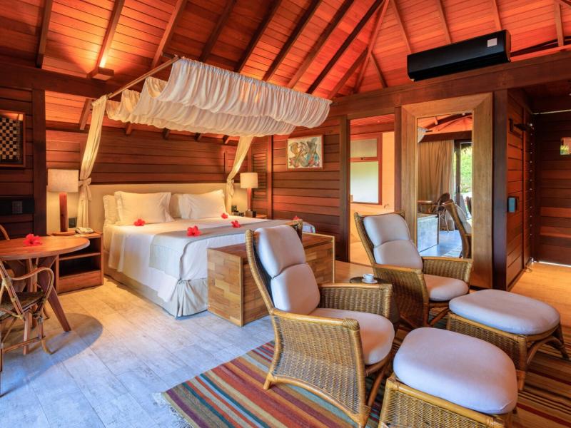 Cozy Garden Bungalow interior featuring a canopy bed and wicker seating, accented by warm reddish-brown walls and soft beige furnishings.