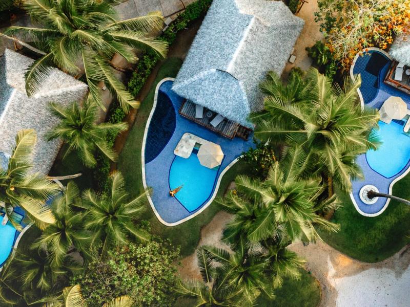 Overhead view of  NANNAI Muro Alto private bungalows with curvy blue pools nestled among rich green palm foliage, accented by natural gray rooftops and sunlit terraces.