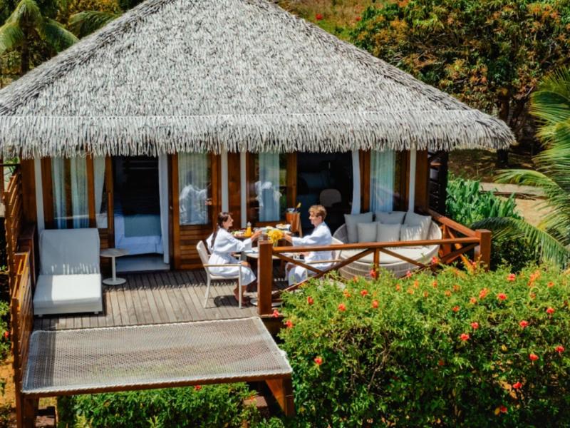 Sea Front Bungalow offers a wooden deck surrounded by lush greenery and vibrant orange blossoms, with cozy white seating under a thatched roof.