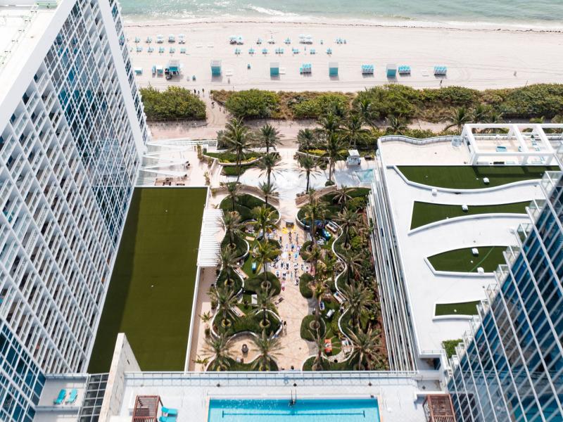 Carillon Miami Wellness Resort expansive aerial view layout featuring a sparkling blue pool, manicured green lawns, and elegant white terraces overlooking the beach.