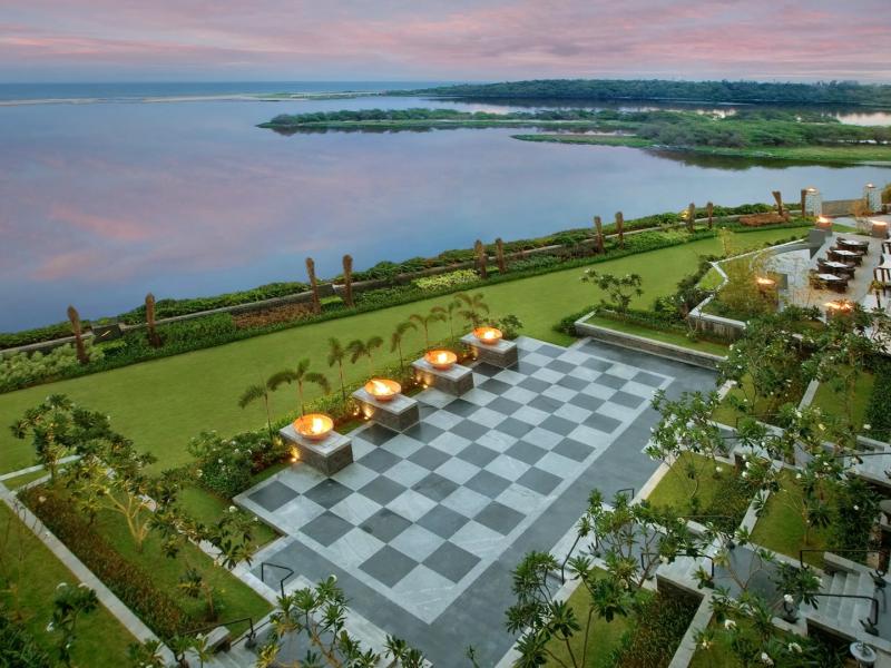 The Leela Palace Chennai seaside façade with lush green lawns and a striking black-and-white checkered courtyard accented by warm golden fire features.