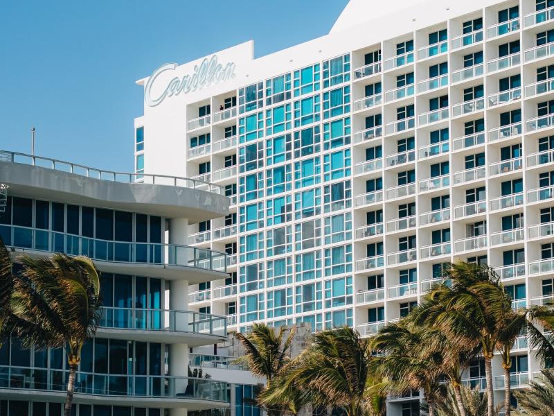 Modern exterior of Carillon Miami Wellness Resort with vibrant turquoise glass accents and balconies framed by lush green palm trees.