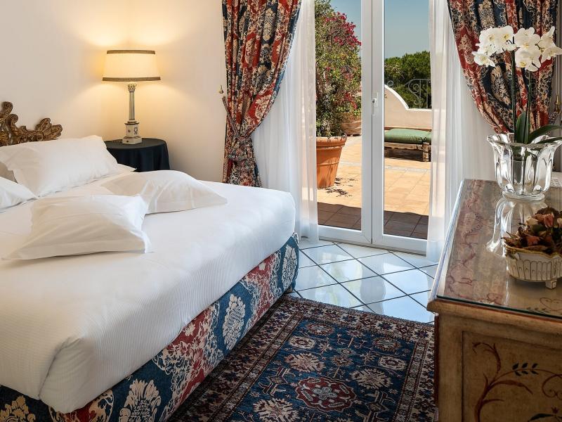Excelsior Belvedere Hotel & SPA Grand Suite bedroom featuring a plush white bed and classic wooden accents, highlighted by rich red-and-blue patterned drapes and a deep navy rug.