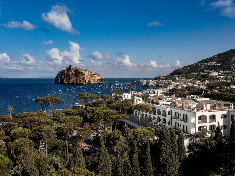 Excelsior Belvedere Hotel & SPA nestled among lush green pines, set against vibrant blue seas and rugged brown cliffs.