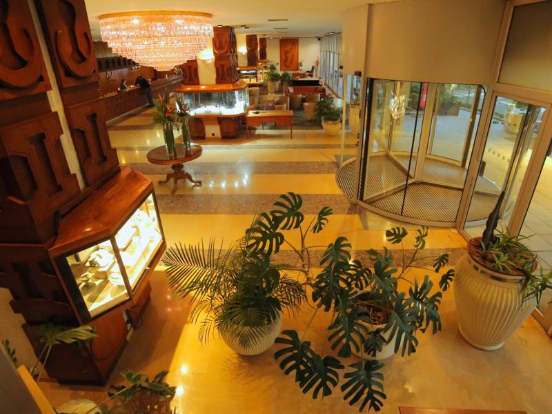 Hotel Carlton warm, golden‑lit lobby entrance welcomes guests with lush green plants, elegant wood accents, and a sparkling chandelier that adds a touch of luxury.