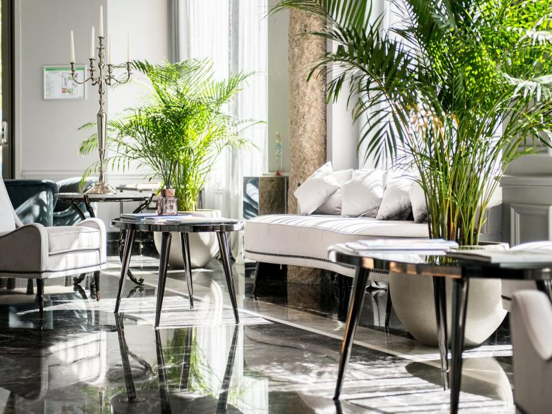 The sunlit lobby lounge blends sleek dark marble floors with soft grey furnishings, enhanced by vibrant green plants and subtle silver décor accents.