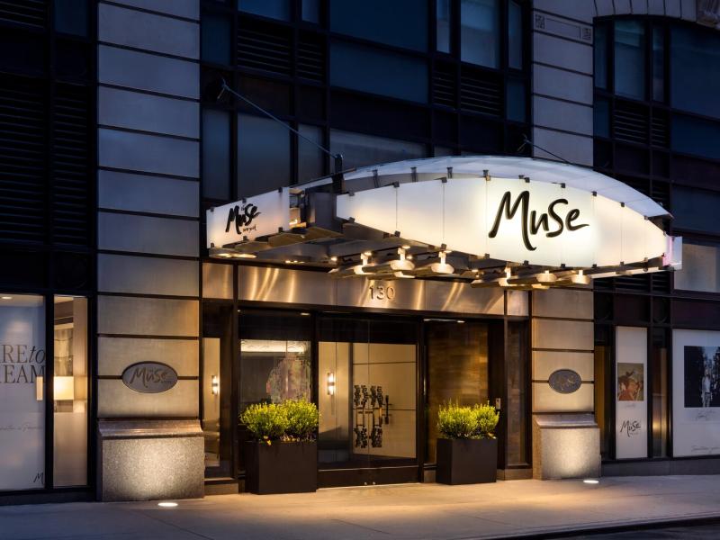 The Muse New York exterior with it’s glowing white canopy accented with warm gold lighting and sleek black metal details welcomes guests into a modern façade framed by soft gray stone.