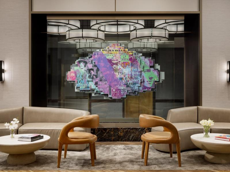 Curved taupe lounge chairs, soft gray carpeting, and elegant bronze lighting in the The Muse New York lobby create a serene seating area centered around a lush floral arrangement.