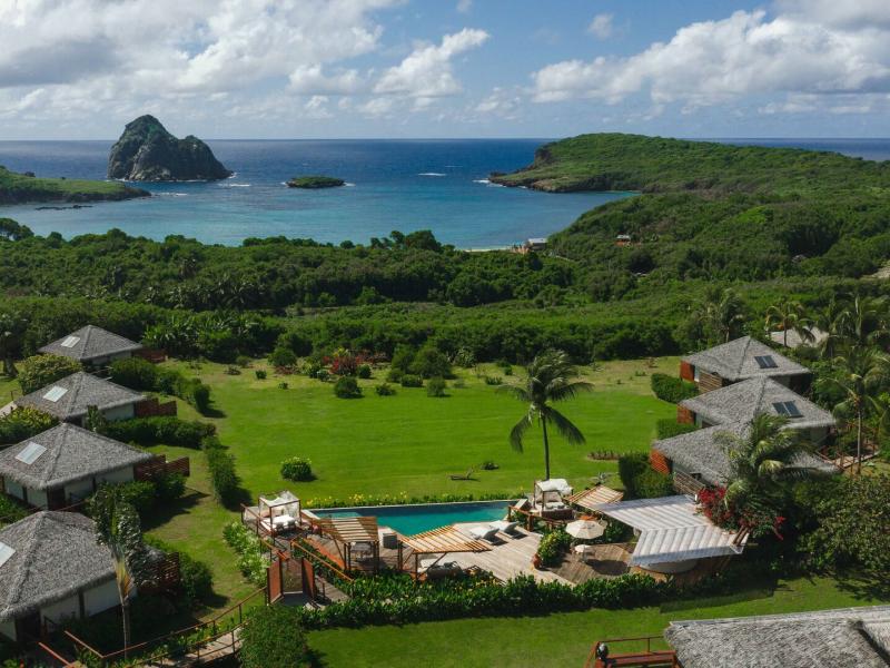 NANNAI Noronha aerial view with breathtaking ocean vistas framed by lush green hills and vibrant turquoise waters, and charming thatched villas and a sparkling pool accented by natural wood tones.