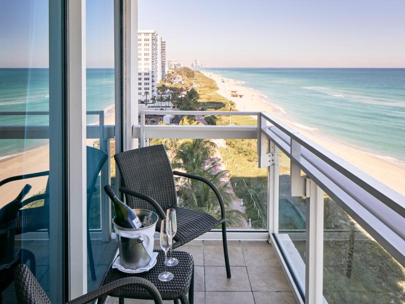 Carillon Miami Wellness Resort private suite balcony with sleek glass railings and dark wicker seating offering breathtaking views of turquoise waters and golden sands.