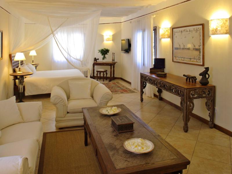 A suite featuring a romantic white canopy bed, plush cream seating, and intricately carved wooden accents set against warm beige walls and soft ambient lighting.