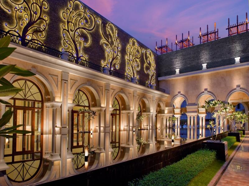 The Leela Palace Chennai Tree of Life Courtyard featuring ornate golden wall designs, arched windows, and shimmering water reflections under a purple twilight sky.