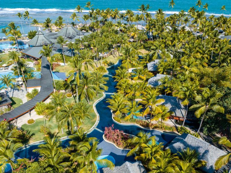 The vibrant resort landscape of NANNAI Muro Alto featuring winding blue pathways and sparkling pools surrounded by dense green palms, set against the bright turquoise sea.
