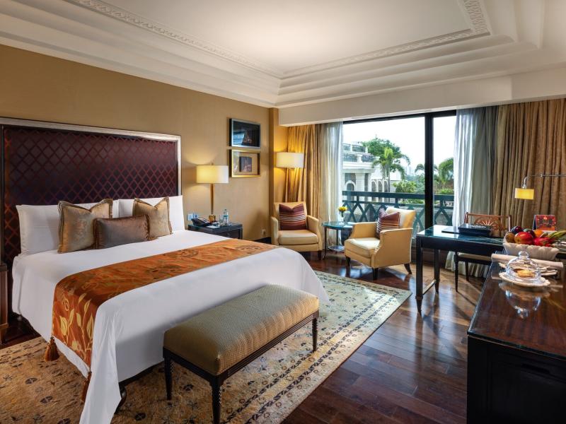 A Royal Club Suite bedroom with city views offers a spacious retreat with rich hardwood floors, warm bronze and gold tones, and elegant seating by the window.