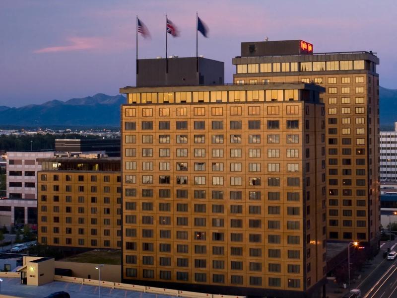The striking city-center Hotel Captain Cook rises above the skyline with a classic high-rise façade, illuminated windows, and rooftop flagpoles, accented by warm golden lighting, soft tan masonry, and deep blue dusk tones.