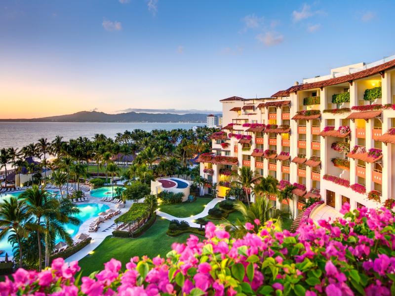 Grand Velas Riviera Nayarit oceanfront resort exterior scene with palm-lined pools and lush gardens, accented by bright turquoise water and vivid pink bougainvillea against warm cream-and-terracotta balconies.