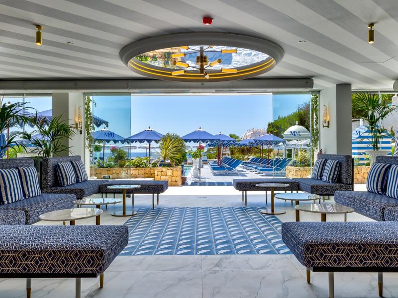 Mongibello Ibiza’s open, airy lobby lounge showcases plush patterned seating, symmetrical layouts, and ocean-view sightlines, accented with navy and white upholstery, brass lighting, and fresh green plants.