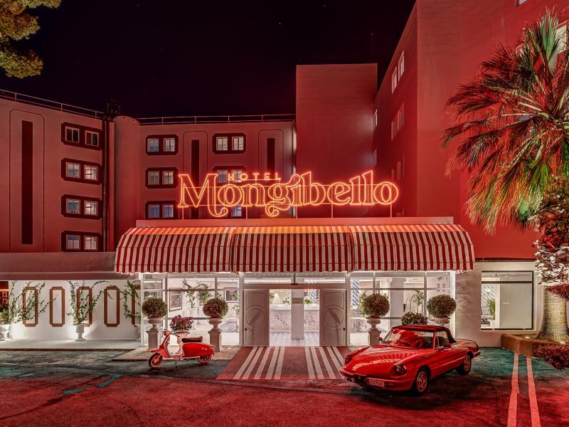 Mongibello Ibiza’s retro-inspired hotel entrance with a striped canopy, palm-lined driveway, and classic car accents, highlighted by bold coral-red architecture with warm white and teal details.