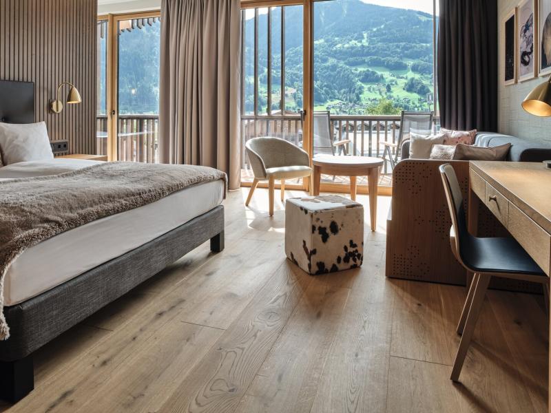 A bright, modern Superior guest room at Loewen Hotel Montafon showcases sweeping mountain views through floor‑to‑ceiling windows, highlighted by light wood furnishings and touches of soft beige and black.