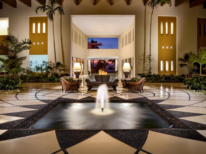 Grand Velas Riviera Nayarit’s grand open-air lobby frames a central fountain and geometric tile floor, highlighted by crisp black-and-white patterns and golden ambient lighting amid rich greenery.