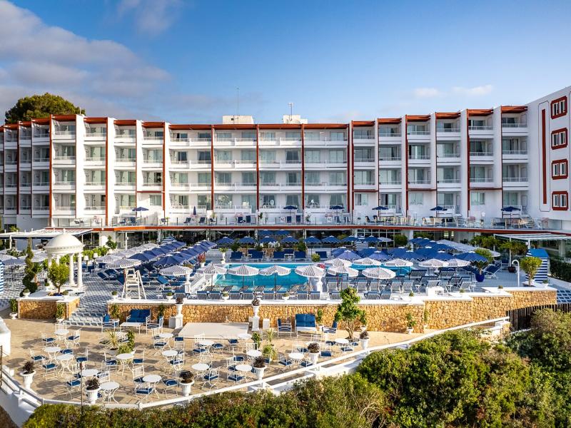 Mongibello Ibiza’s sweeping façade overlooks a terraced pool deck with multiple swimming pools and loungers, accented by crisp white structures, sky-blue water, and sandy beige stone.