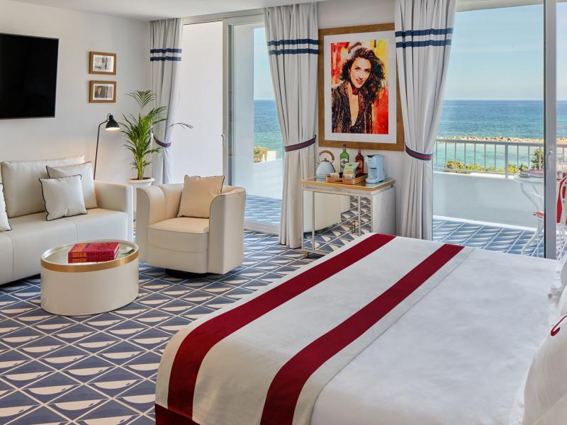 Mongibello Ibiza’s bright, open Sea View Terrace Suite bedroom features a plush king bed, modern lounge seating, and floor-to-ceiling terrace doors, accented with crisp white, navy blue, and bold red details.