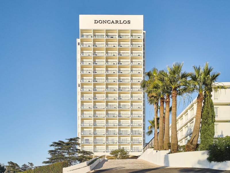 Don Carlos Marbella striking hotel tower rises above palm‑lined grounds with sun‑washed balconies and panoramic views, accented by crisp white architecture, warm sand tones, and a brilliant Mediterranean blue sky.