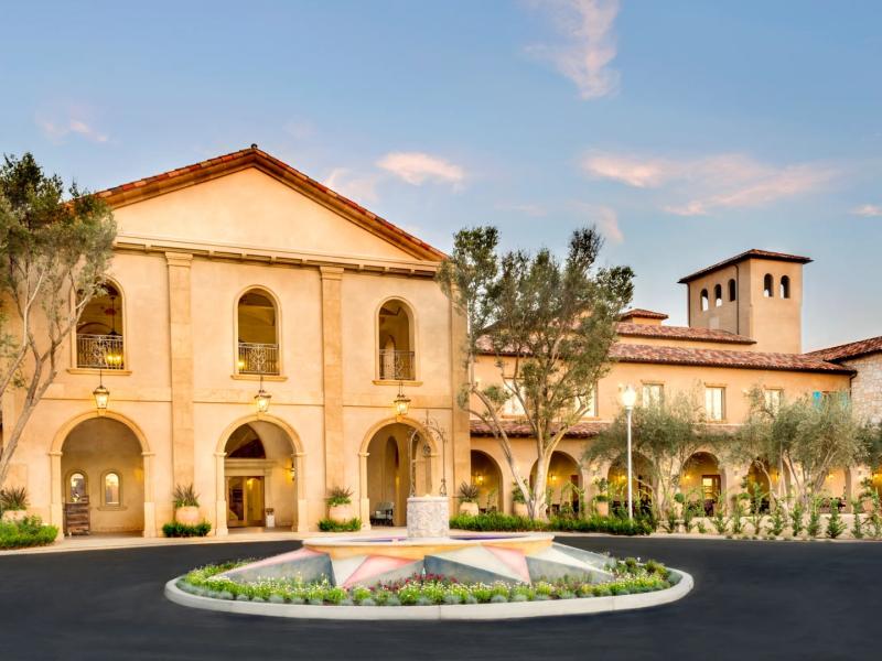 The grand Mediterranean-style entrance of Allegretto Vineyard Resort showcases arched colonnades, a circular drive, and warm stone architecture accented by soft beige, terracotta, and lush green tones.