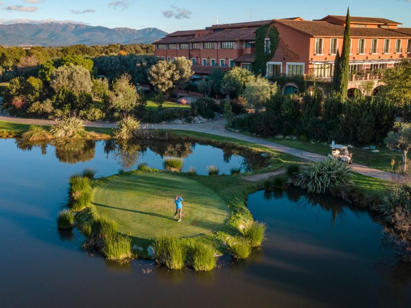 This golf course’s signature island green sits surrounded by shimmering blue water and lush emerald landscaping, framed by Hotel Peralada terracotta‑toned facade and mountain backdrop.