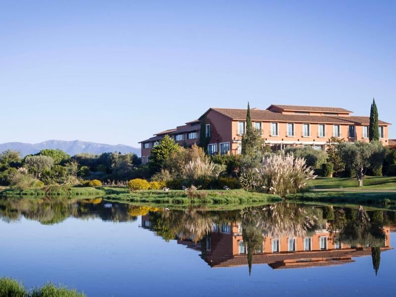 A serene lake adjacent to Hotel Peralada reflects the terracotta‑toned facade and lush green gardens beneath a vibrant blue sky.