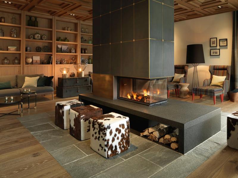 Loewen Hotel Montafon warm, modern lounge centered around a sleek double‑sided fireplace features cowhide-accented stools and built‑in wood shelving, enhanced by rich earthy browns and soft charcoal tones.