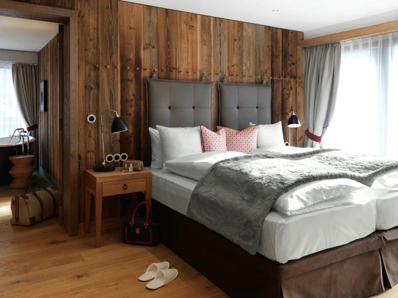 A cozy alpine-style bedroom in a Löwen suite at Loewen Hotel Montafon features a rustic wood accent wall and plush gray bedding, complemented by soft pink pillows and warm natural wood tones.