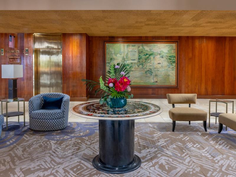 Hotel Captain Cook’s welcoming lobby showcases a round stone table with a floral arrangement, patterned carpeting, and rich wood-paneled walls, accented by deep navy seating, forest green